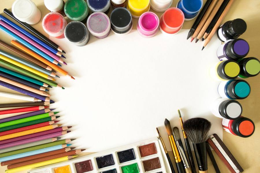 Overhead view of colour pencils, paint jars, brushes and a watercolour set framing a sheet of white paper