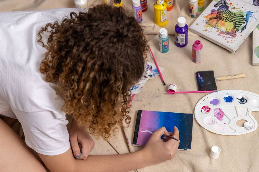 Young artist painting a purple and blue sky on a small canvas, surrounded by acrylic paint bottles and a palette