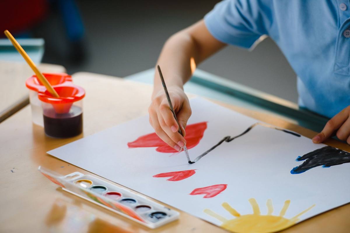 Child’s hands painting red hearts and a yellow sun with watercolours at a classroom table