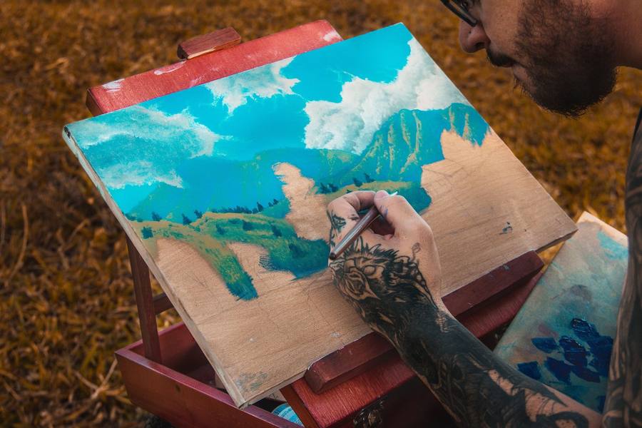 Artist painting a bright landscape of mountains, clouds and hills on a wooden easel outdoors