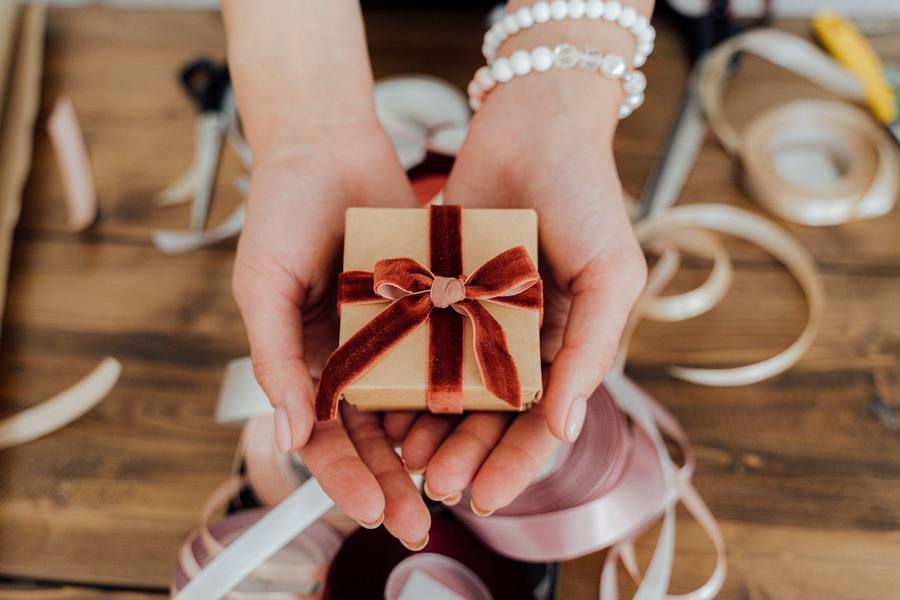 Hands holding a small kraft-paper gift box tied with a red velvet bow, ribbons on the table