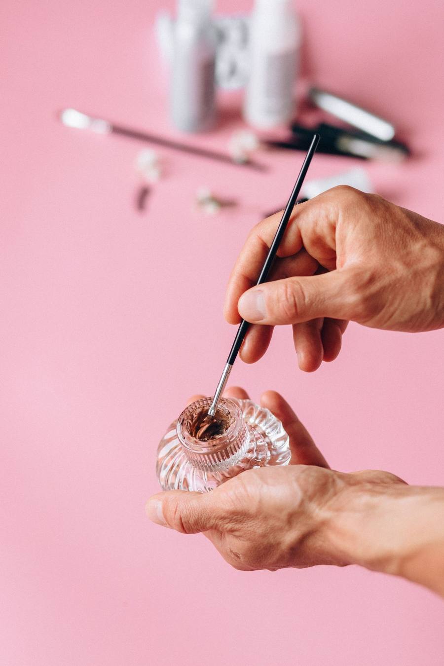 Hands painting a small glass bottle with a fine brush against a pink background