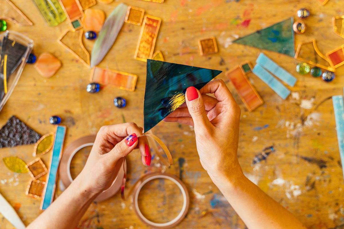 Hands arranging colourful glass and paper pieces on a paint-splattered craft table