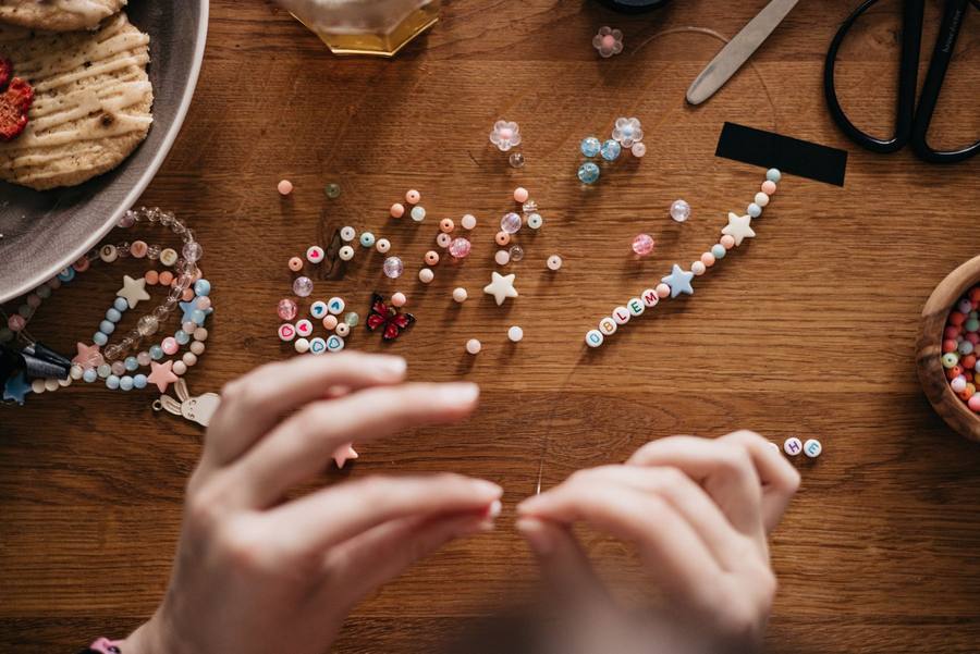 Hands stringing letter and star beads into bracelets on a wooden table with craft tools