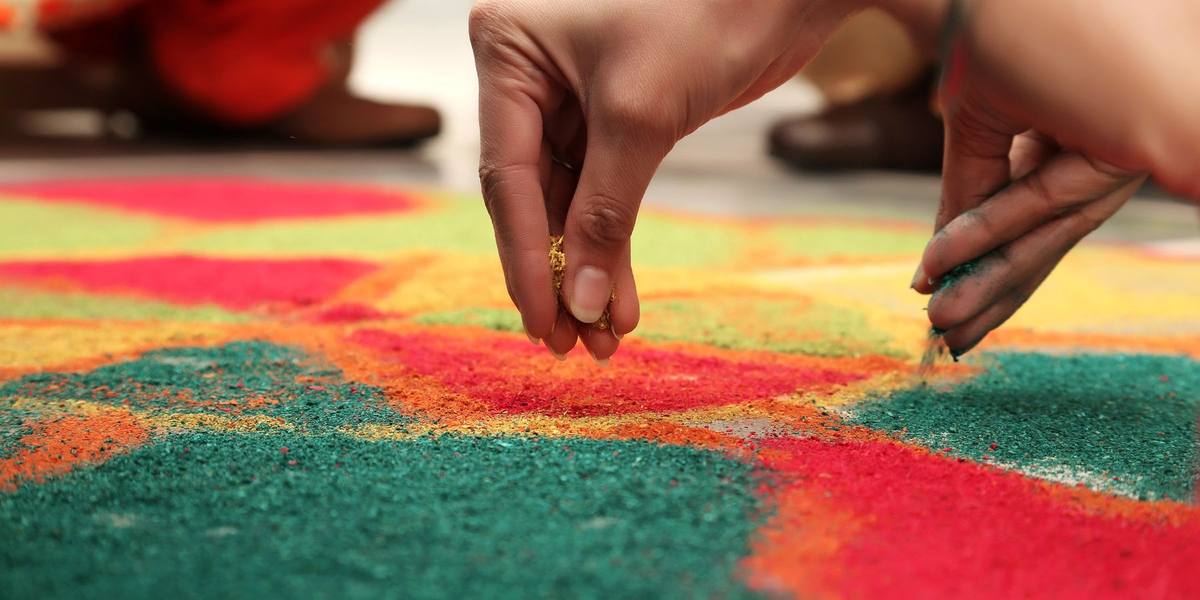 Hands sprinkling coloured powder to fill a red, green and yellow rangoli design