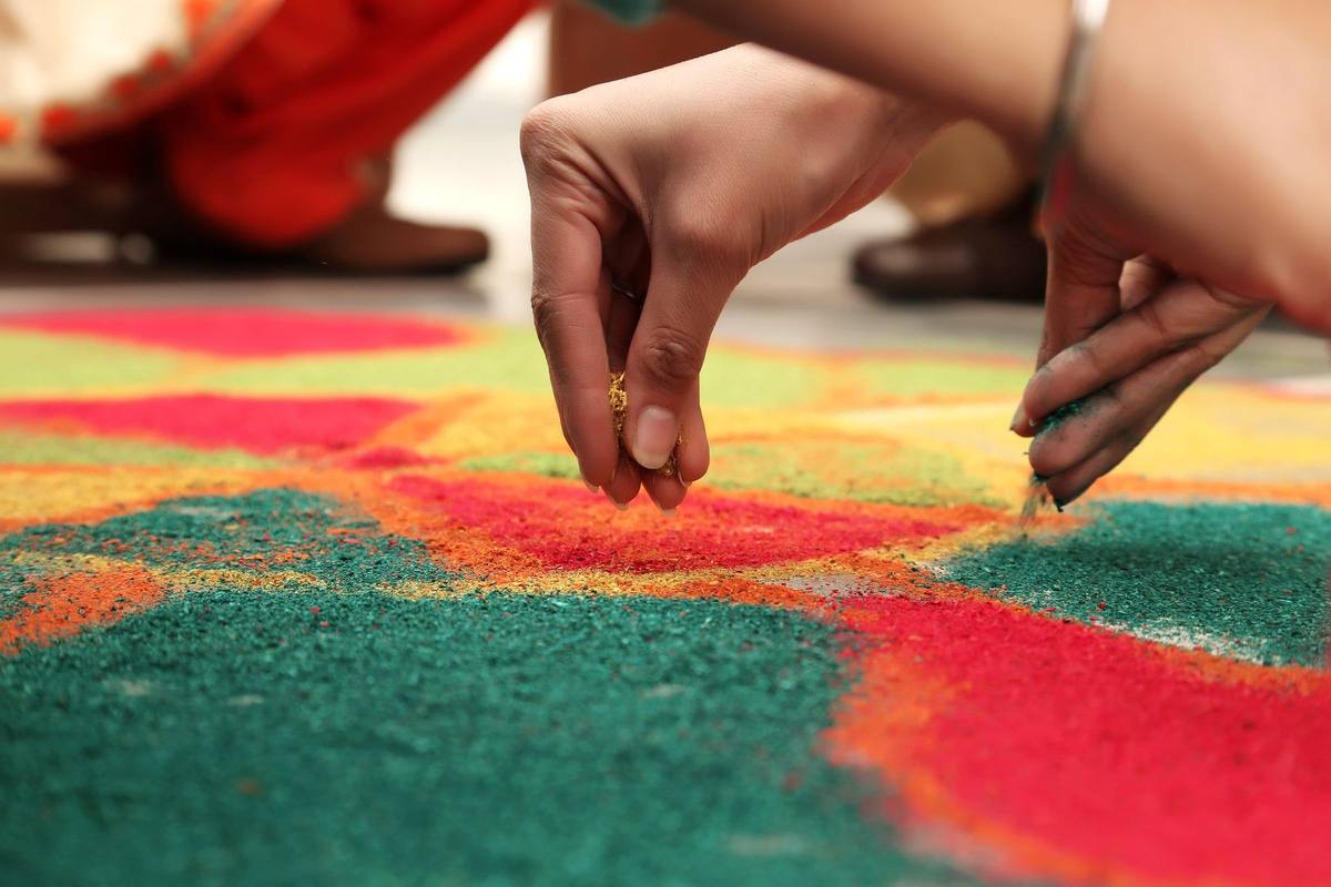 Hands sprinkling coloured powder to fill a red, green and yellow rangoli design