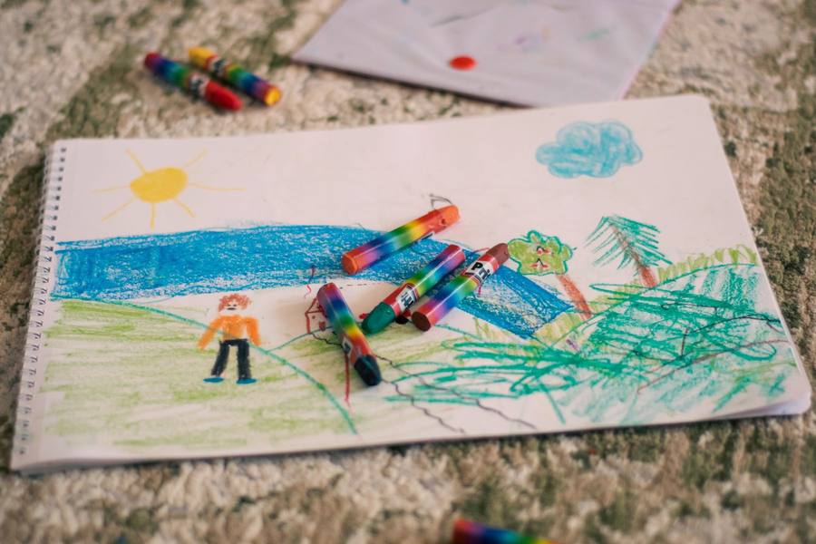 Child’s crayon drawing of a sunny landscape with a river, trees and a figure, crayons resting on top